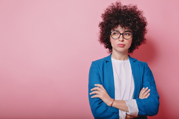 Mujer de negocios moderna vestida con chaqueta azul y usa gafas posa en rosa con emociones insatisfechas