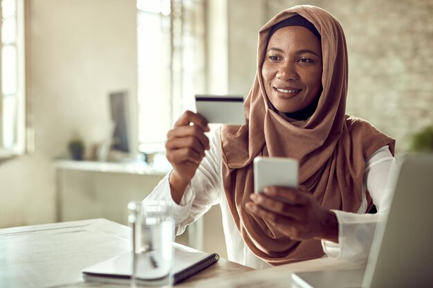 Mujer de negocios musulmana sonriente comprando en línea mientras usa tarjeta de crédito y teléfono móvil en la oficina