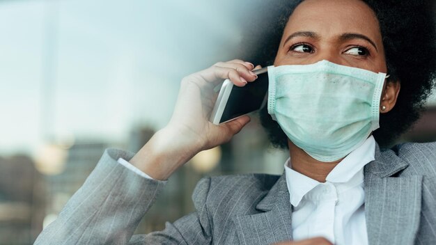 Mujer de negocios negra angustiada que se comunica por teléfono inteligente mientras usa una máscara facial durante la epidemia de virus La vista es a través del cristal