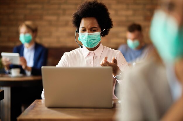 Mujer de negocios negra con mascarilla protectora haciendo videollamadas a través de una laptop en un café