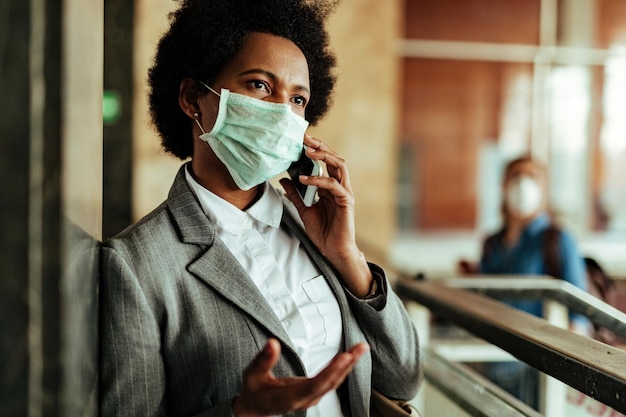 Mujer de negocios negra con mascarilla protectora mientras se comunica por teléfono móvil en el corredor de la estación de tren