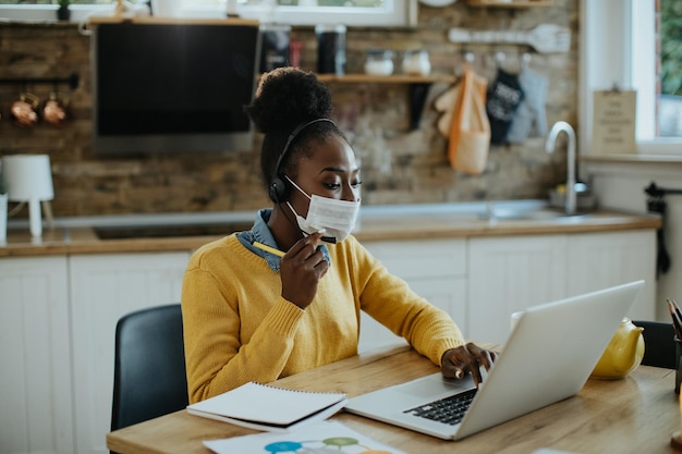 Mujer de negocios negra que usa computadora mientras realiza una llamada de conferencia en casa durante la pandemia de coronavirus