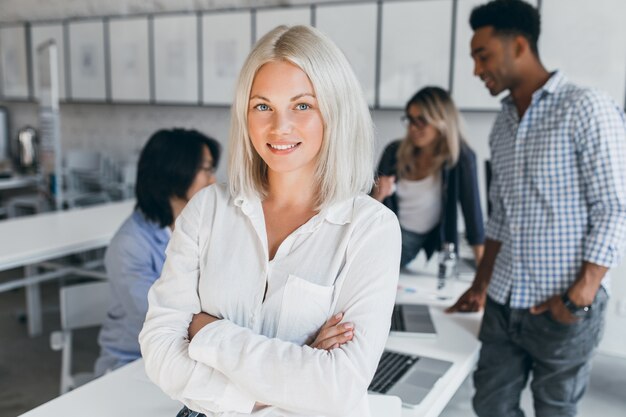 Mujer de negocios de ojos azules en blusa blanca de pie en pose de confianza con sus compañeros de trabajo internacionales. Retrato interior de empleados asiáticos y africanos con dama rubia.