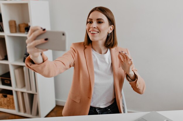 Mujer de negocios de ojos marrones con chaqueta de color melocotón se hace selfie en su nueva oficina blanca