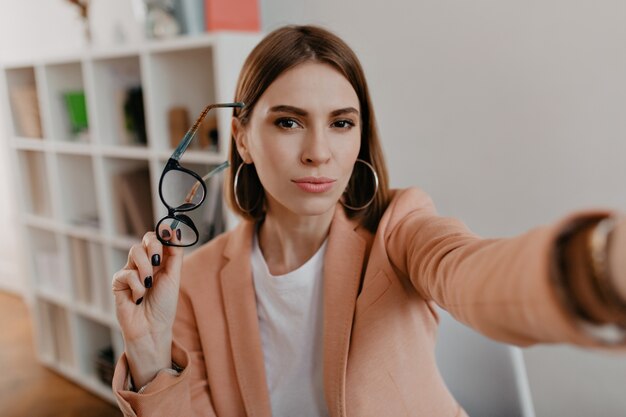 Mujer de negocios de ojos marrones se quitó las gafas y se toma un selfie en su oficina blanca.