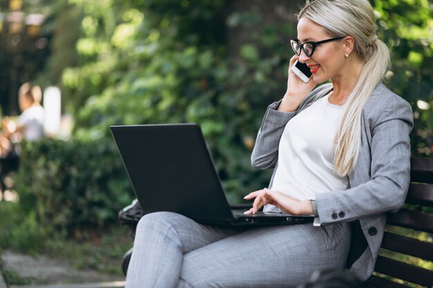 Mujer de negocios con ordenador portátil hablando por teléfono en el parque en un banco