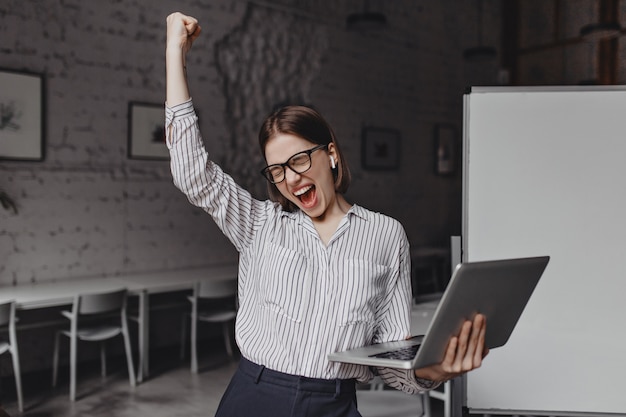 Mujer de negocios con ordenador portátil en mano está feliz con el éxito. Retrato de mujer con gafas y blusa a rayas gritando con entusiasmo y haciendo gesto ganador.