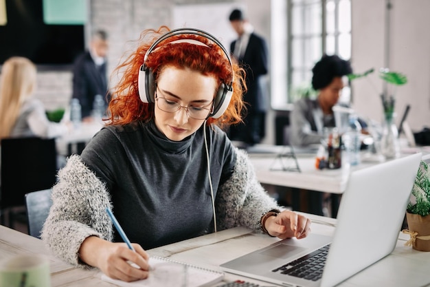 Mujer de negocios pelirroja que usa una computadora portátil y escribe notas en su bloc de notas mientras escucha música en los auriculares en el trabajo