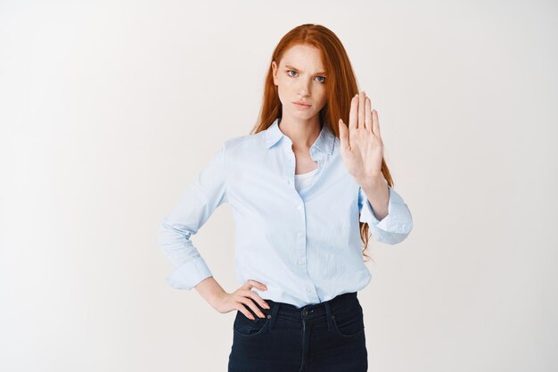 Mujer de negocios pelirroja seria mostrando señal de stop y frunciendo el ceño disgustado, diciendo que no, negándose a algo