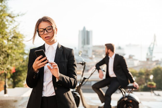 Mujer de negocios pensativa que presenta al aire libre y que usa el teléfono