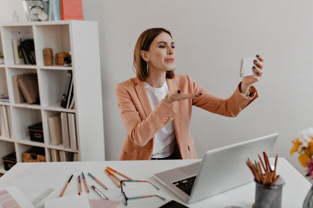 Mujer de negocios positiva con traje elegante envía beso de aire a la cámara del teléfono mientras está sentado en el lugar de trabajo.