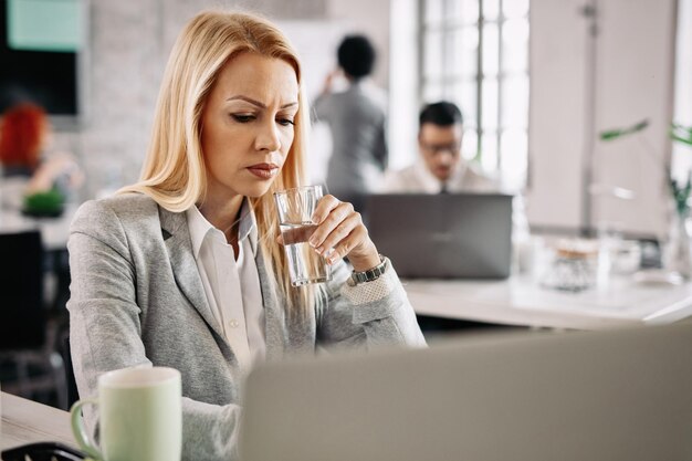 Mujer de negocios preocupada que trabaja en una computadora mientras bebe agua de un vaso en la oficina