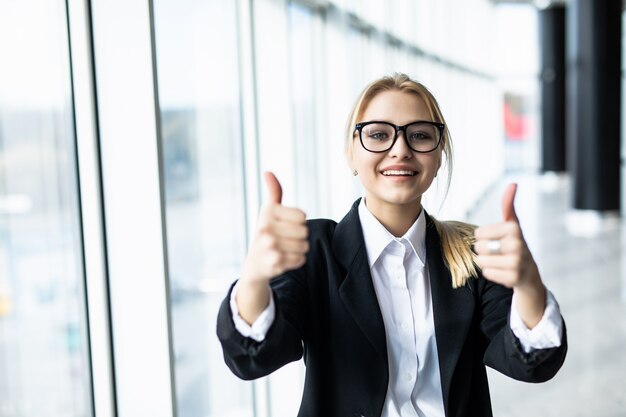 Mujer de negocios con los pulgares para arriba en la oficina con ventanas panorámicas