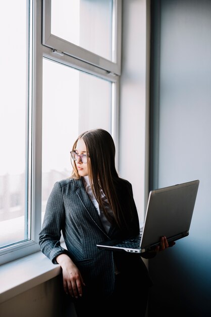 Mujer de negocios que se coloca en la ventana con la computadora portátil