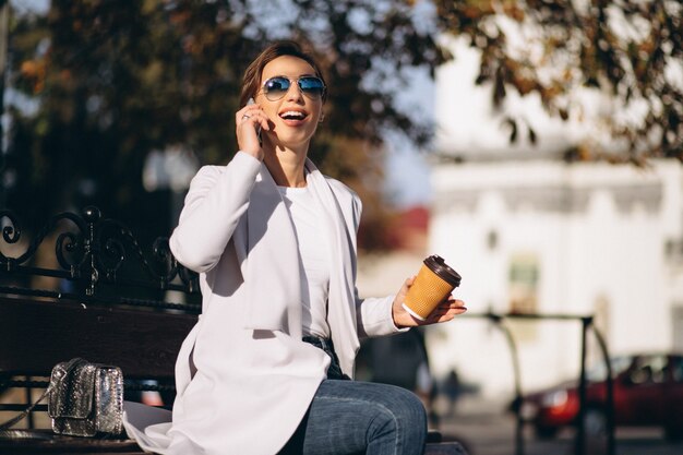 Mujer de negocios que se sienta en un banco con café de consumición del teléfono