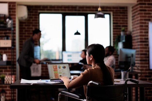 Mujer de negocios que vive con discapacidad analizando el informe en una computadora portátil, trabajando con estadísticas en una oficina apta para discapacitados. Empleado asiático que sufre de discapacidad crónica, usa silla de ruedas.