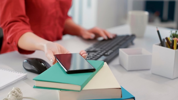 Mujer de negocios revisando el teléfono móvil en el escritorio en el trabajo de oficina, mirando el mensaje de texto. Empleada que trabaja en desarrollo financiero con estadísticas de ventas en computadora. Disparo de mano. De cerca.