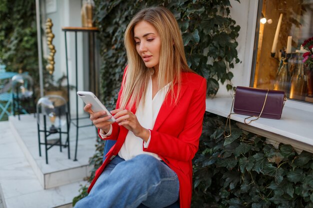 Mujer de negocios rubia confiada en chaqueta elegante roja usando teléfono mobyle y sentado en el café de la ciudad