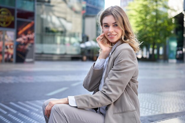 Mujer de negocios rubia confiada en traje sonríe a la cámara se sienta al aire libre cerca de edificios de oficinas