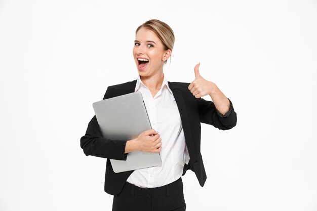 Mujer de negocios rubia feliz sosteniendo la computadora portátil y mostrando el pulgar hacia arriba mientras que sobre la pared blanca