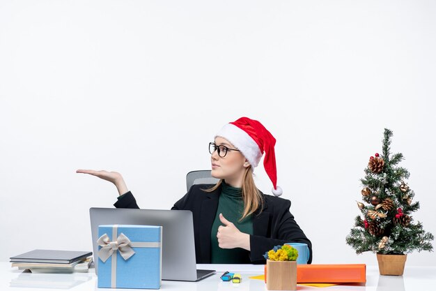 Mujer de negocios rubia con sombrero de santa claus sentado en una mesa con un árbol de Navidad y un regalo en él haciendo un gesto de ok y apuntando algo en el lado derecho de la oficina sobre fondo blanco