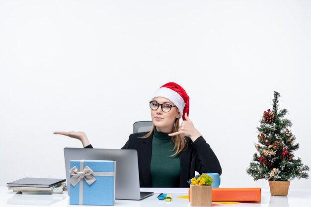 Mujer de negocios rubia con sombrero de santa claus sentado en una mesa con un árbol de Navidad y un regalo haciendo gesto de llamarme y señalando algo en el lado derecho en la oficina sobre fondo blanco