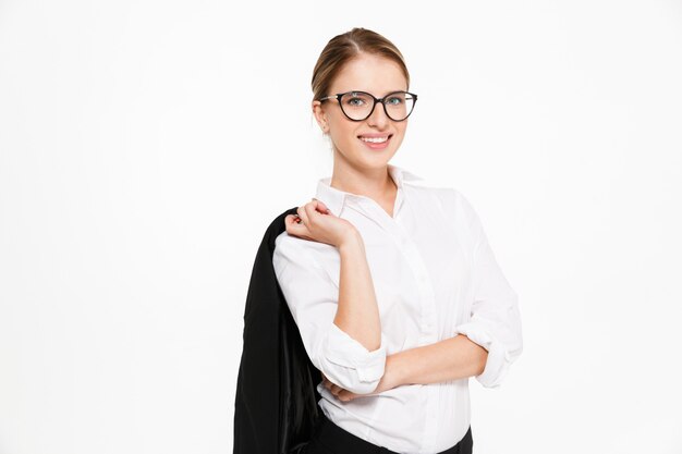 Mujer de negocios rubia sonriente en anteojos sosteniendo su chaqueta y sobre la pared blanca