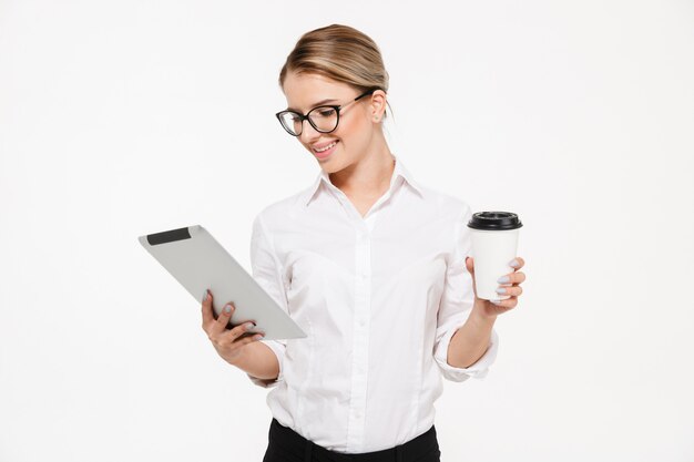 Mujer de negocios rubia sonriente en anteojos usando tableta mientras sostiene la taza de té sobre la pared blanca