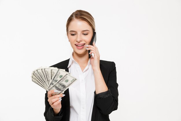 Mujer de negocios rubia sonriente hablando por los teléfonos mientras sostiene y mira dinero sobre la pared blanca