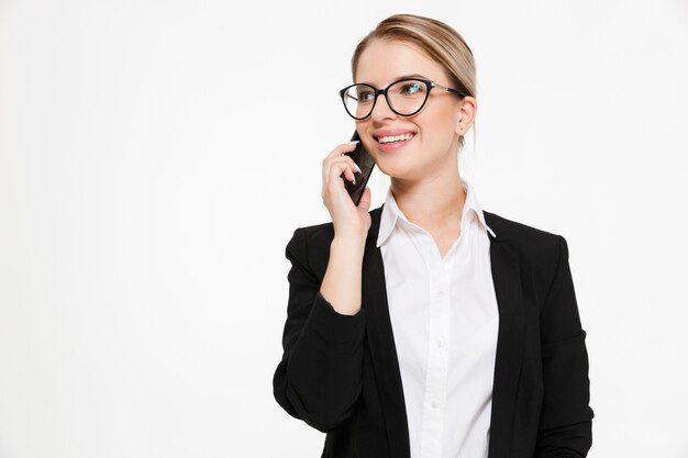 Mujer de negocios rubia sonriente en lentes que habla por el teléfono y que mira lejos sobre blanco