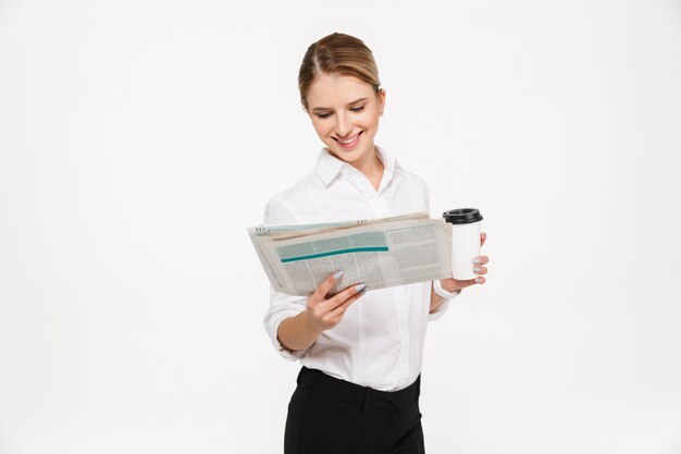 Mujer de negocios rubia sonriente leyendo el periódico mientras sostiene la taza de café sobre la pared blanca
