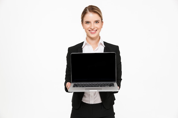 Mujer de negocios rubia sonriente que muestra la pantalla de la computadora portátil en blanco y sobre la pared blanca