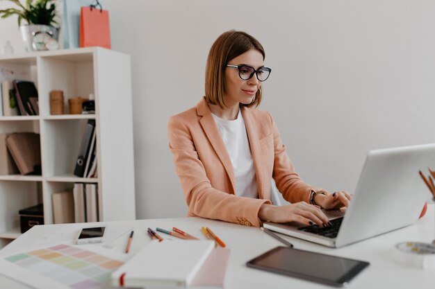 Mujer de negocios satisfecha con gafas trabajando en equipo portátil. Retrato de mujer joven en traje elegante de muebles de oficina.