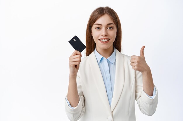 Mujer de negocios segura y exitosa muestra tarjeta de crédito de plástico y pulgares hacia arriba, sonriendo complacida, recomienda banco, de pie sobre una pared blanca