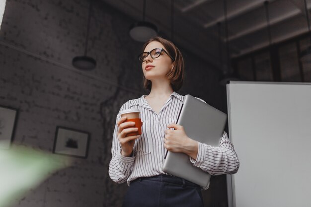 Mujer de negocios seria en blusa elegante y pantalón negro sosteniendo portátil y tomando café en la oficina.