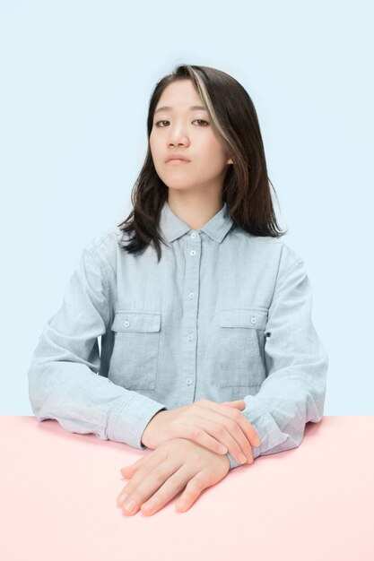 Mujer de negocios seria sentada en la mesa, mirando a cámara aislada sobre fondo azul de moda estudio. Rostro joven y hermoso.