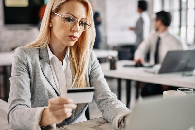 Una mujer de negocios seria con una tarjeta de crédito y una computadora portátil para comprar en línea mientras está sentada en el escritorio de su oficina