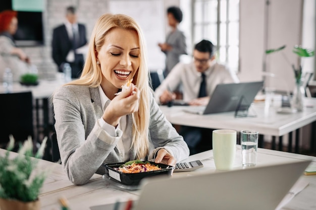 Mujer de negocios sonriente comiendo alimentos saludables en el trabajo y comiendo ensalada de verduras para el almuerzo