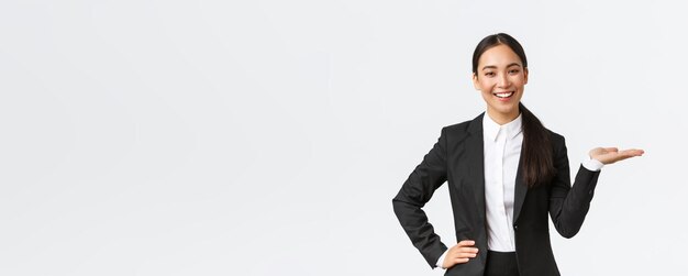 Una mujer de negocios sonriente profesional presenta su proyecto durante una reunión con una vendedora de traje negro sosteniendo la mano a la derecha mientras muestra el producto sosteniendo la palma sobre un fondo blanco en blanco