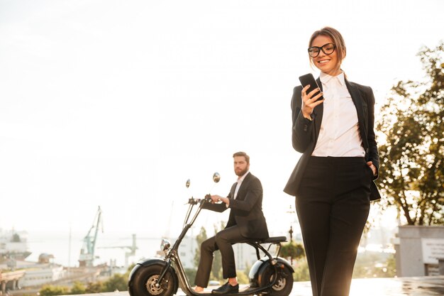 Mujer de negocios sonriente que presenta al aire libre y que usa el teléfono