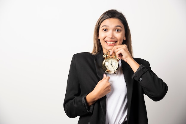 Mujer de negocios sosteniendo un reloj de alarma en la pared blanca.