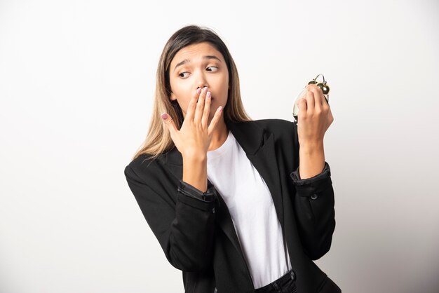 Mujer de negocios sosteniendo un reloj de alarma en la pared blanca.