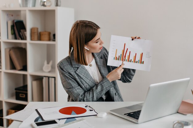 La mujer de negocios sostiene el gráfico en sus manos y se lo cuenta a los clientes a través de la comunicación por video mientras está sentada en un lugar de trabajo brillante.