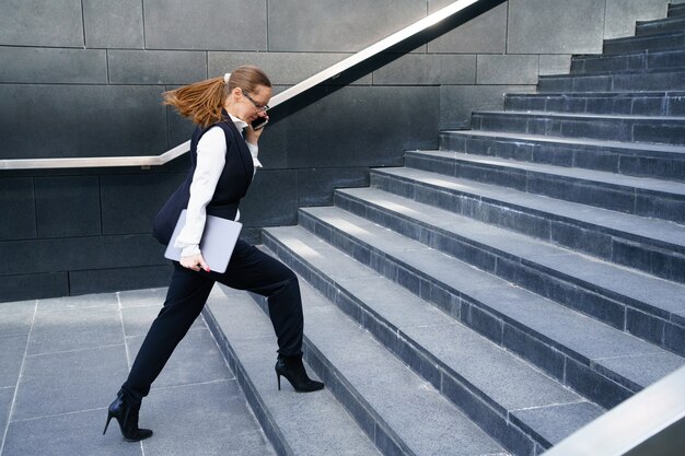 Una mujer de negocios sube las escaleras sosteniendo una tableta en la mano y hablando por teléfono.