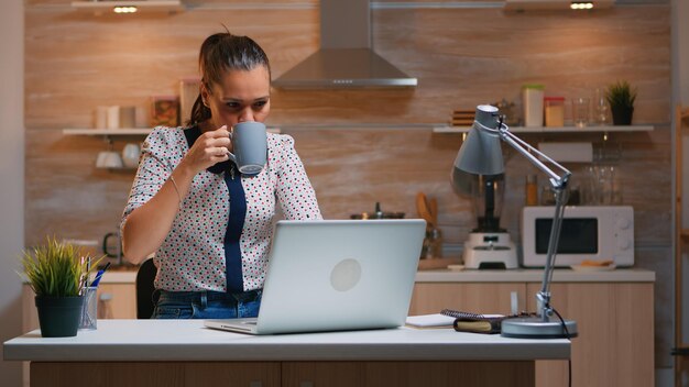 Mujer de negocios trabajando desde casa por la noche escribiendo en la computadora portátil y tomando café. Empleado enfocado ocupado que usa la red inalámbrica de tecnología moderna haciendo horas extras para leer el trabajo, escribir, buscar