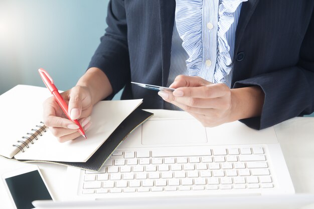 Mujer de negocios en traje oscuro explotación de tarjeta de crédito, utilizando equipo portátil y escritura en el bloc de notas, Negocios y concepto de compras en línea