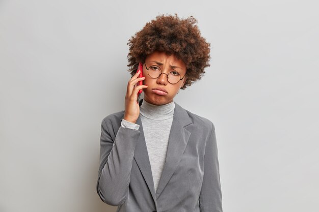 Mujer de negocios triste agotada con cabello rizado, habla por teléfono, vestida con un traje formal de moda, tiene una conversación aburrida, se ve triste, suspira de cansancio