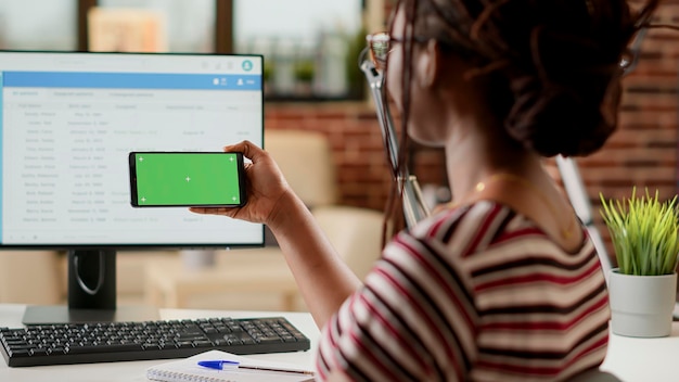 Mujer de negocios usando pantalla verde horizontal en el teléfono inteligente, sentada en el escritorio. Freelancer mirando el fondo de la maqueta en blanco con copyspace aislado y plantilla de clave de croma en la pantalla del teléfono.