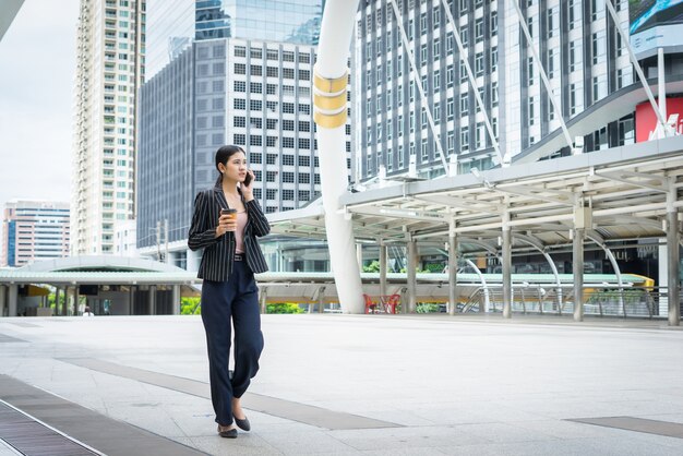 Mujer de negocios usando teléfono con café en la mano caminando en la calle con edificios de oficinas en el fondo