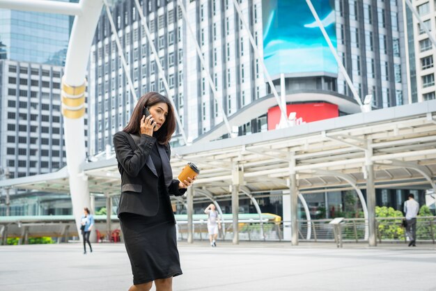 Mujer de negocios usando teléfono con café en la mano caminando en la calle con edificios de oficinas en el fondo